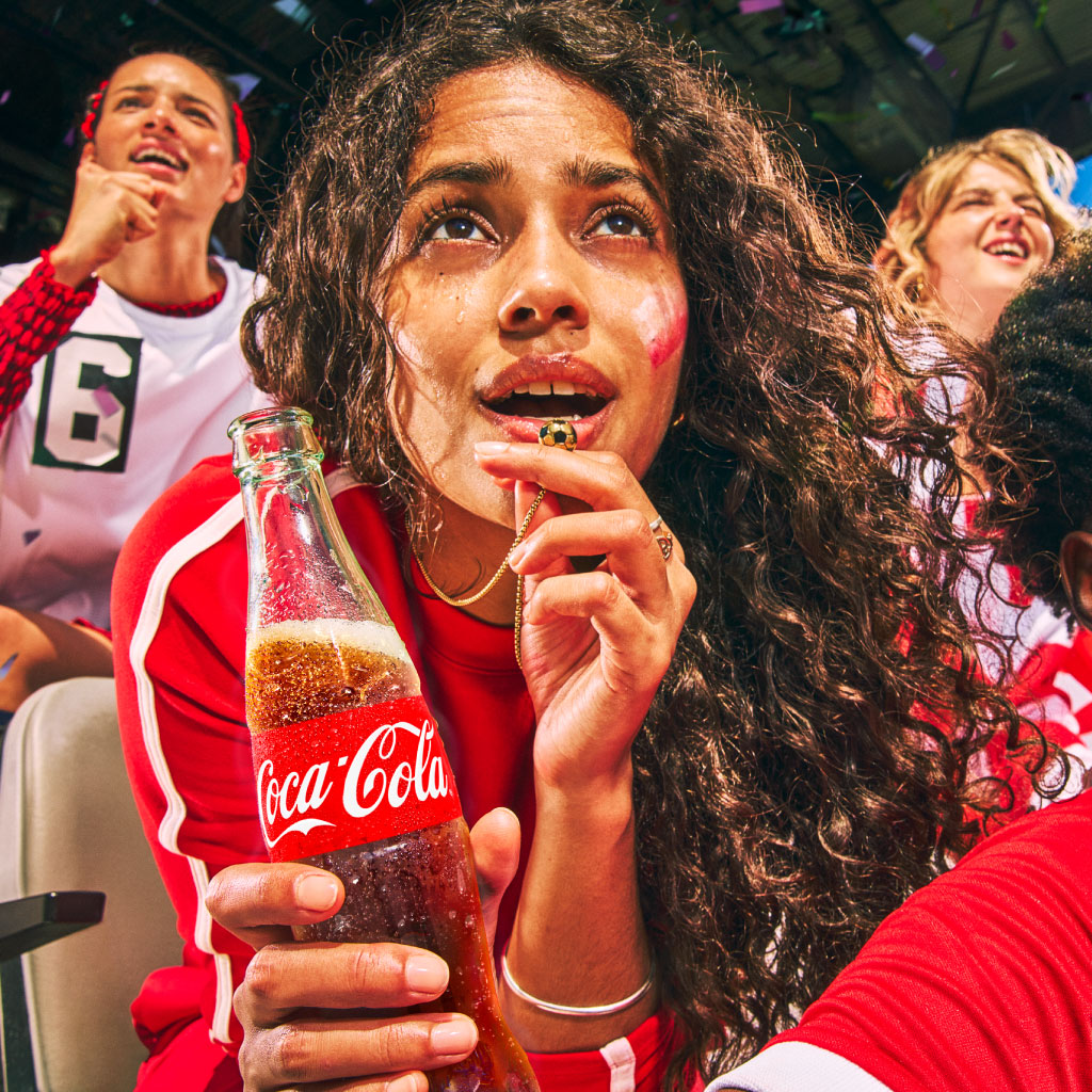 Pessoa a segurar uma garrafa de vidro de Coca‑Cola, rodeada por fãs vestidos com roupas vermelhas num ambiente de estádio.