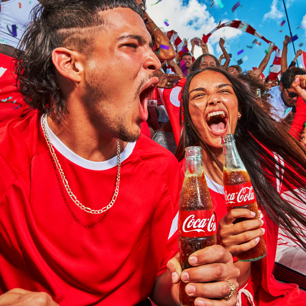 Grupo de pessoas vestidas de vermelho a celebrar ao ar livre, segurando garrafas de Coca‑Cola, com confettis no ar e um ambiente festivo e energético.
