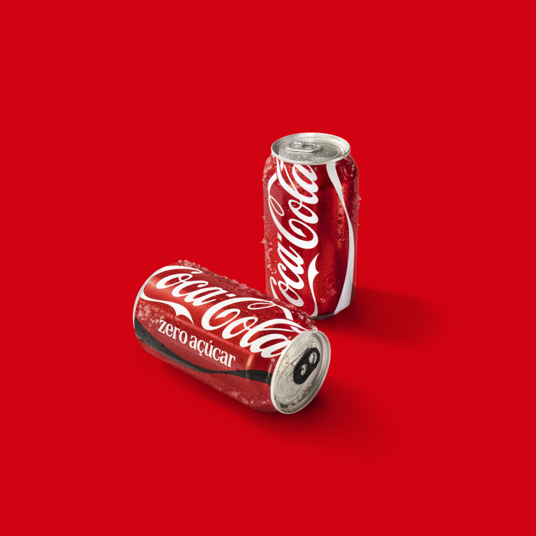 Duas latas de Coca‑Cola sem açúcar sobre fundo vermelho, com gotas de condensação e branding da marca em destaque.