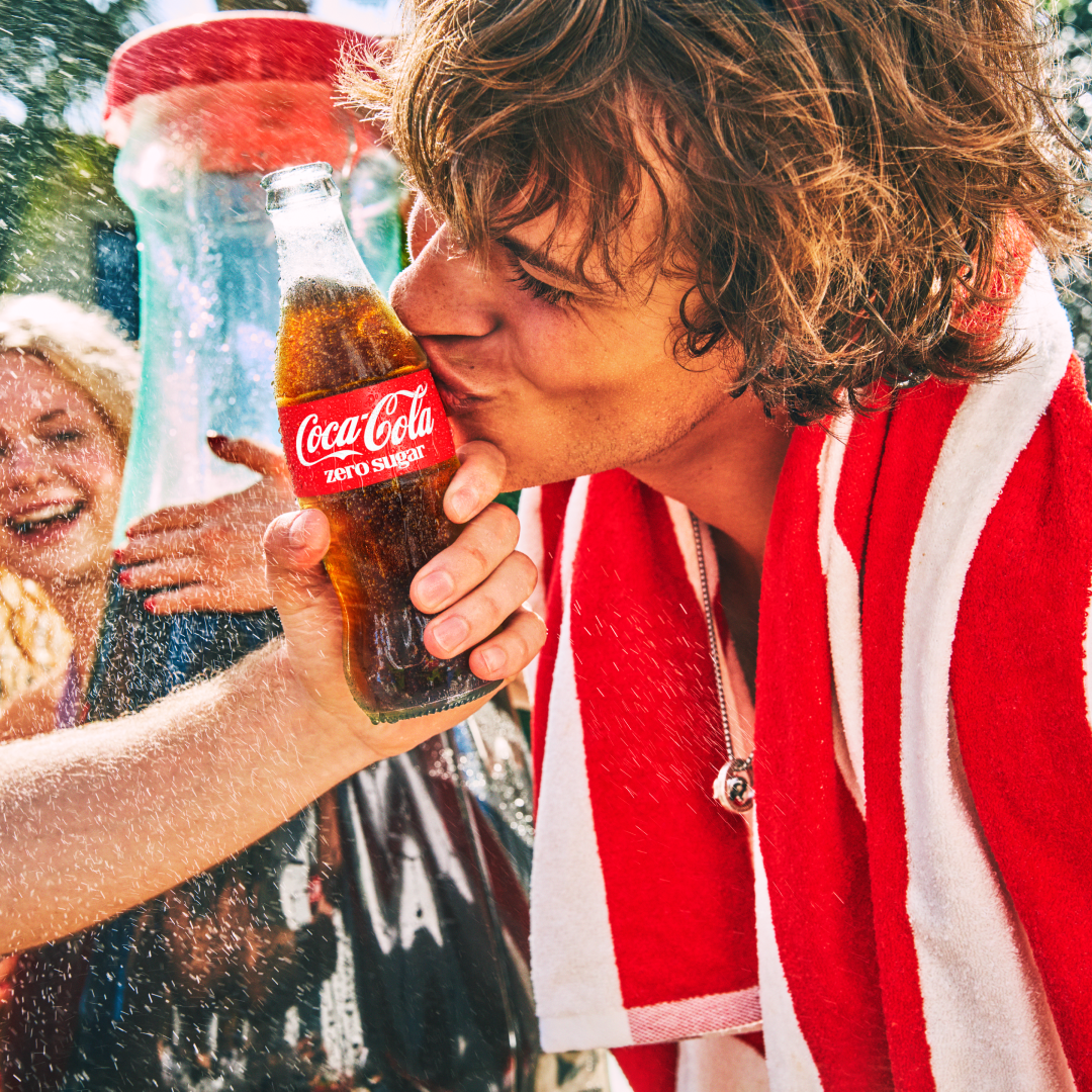 Pessoa enrolada numa toalha vermelha e branca a segurar uma garrafa de Coca‑Cola junto ao rosto, num ambiente de verão e diversão ao ar livre