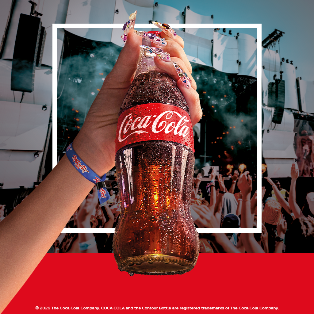 Pessoa a segurar uma garrafa de vidro de Coca‑Cola erguida no ar durante um concerto com o público em festa, evocando a experiência do Rock in Rio Lisboa 2026