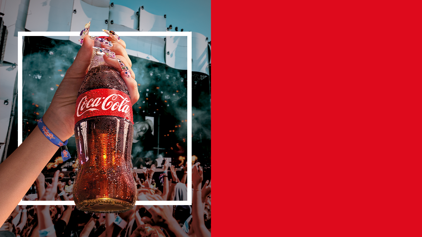 Pessoa a segurar uma garrafa de Coca‑Cola erguida no ar durante um concerto, com o público em festa ao fundo.