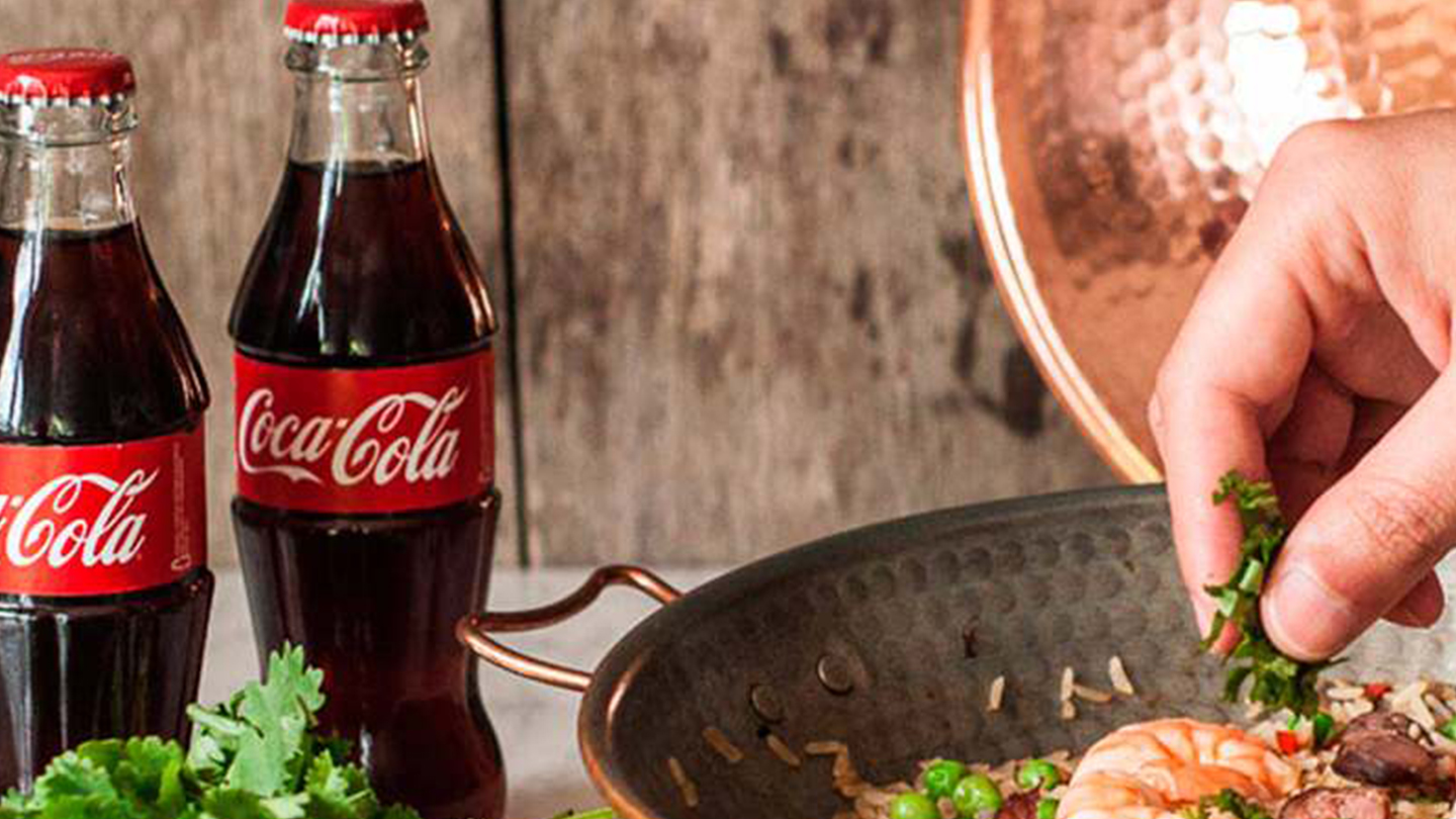 Detalhe de uma mão temperando uma tijela de comida ao lado de duas garrafas de Coca-Cola