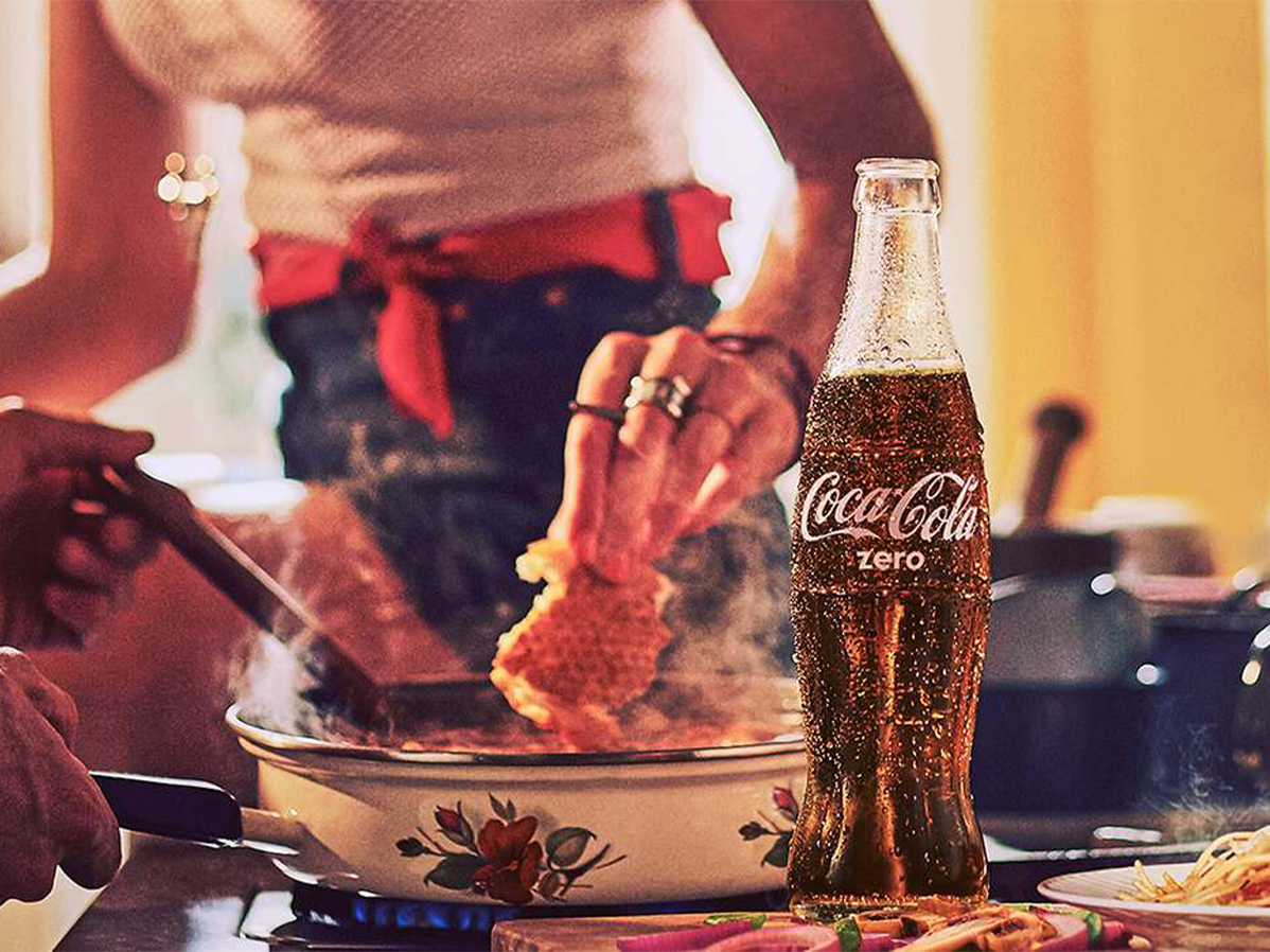 Duas pessoas cozinhando em uma frigideira com uma garrafa de Coca-Cola Zero