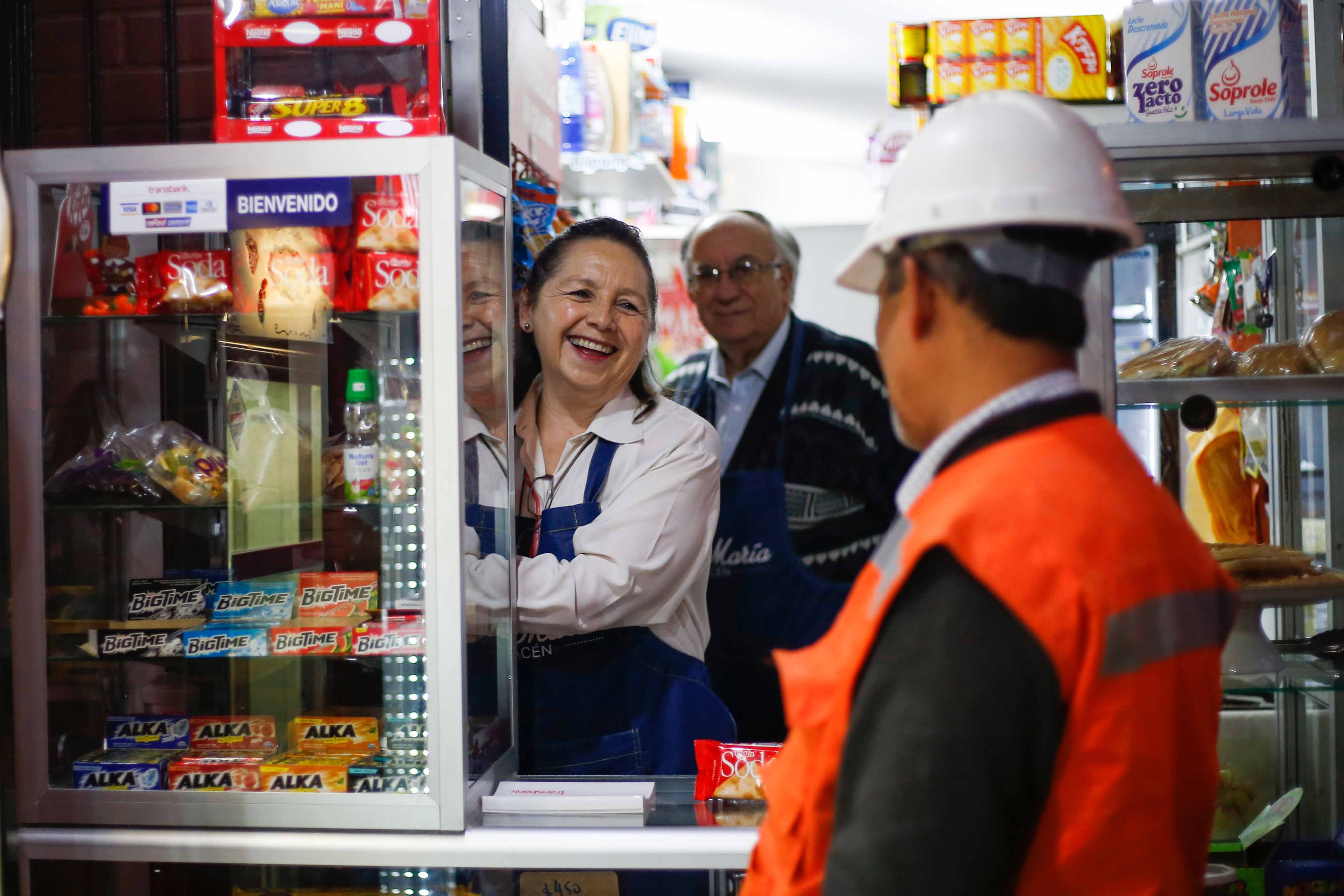 Una mujer atiende a clientes en un pequeño almacén o kiosco. Un hombre con casco de seguridad está de espaldas a la cámara. Se ven estantes con diversos productos.