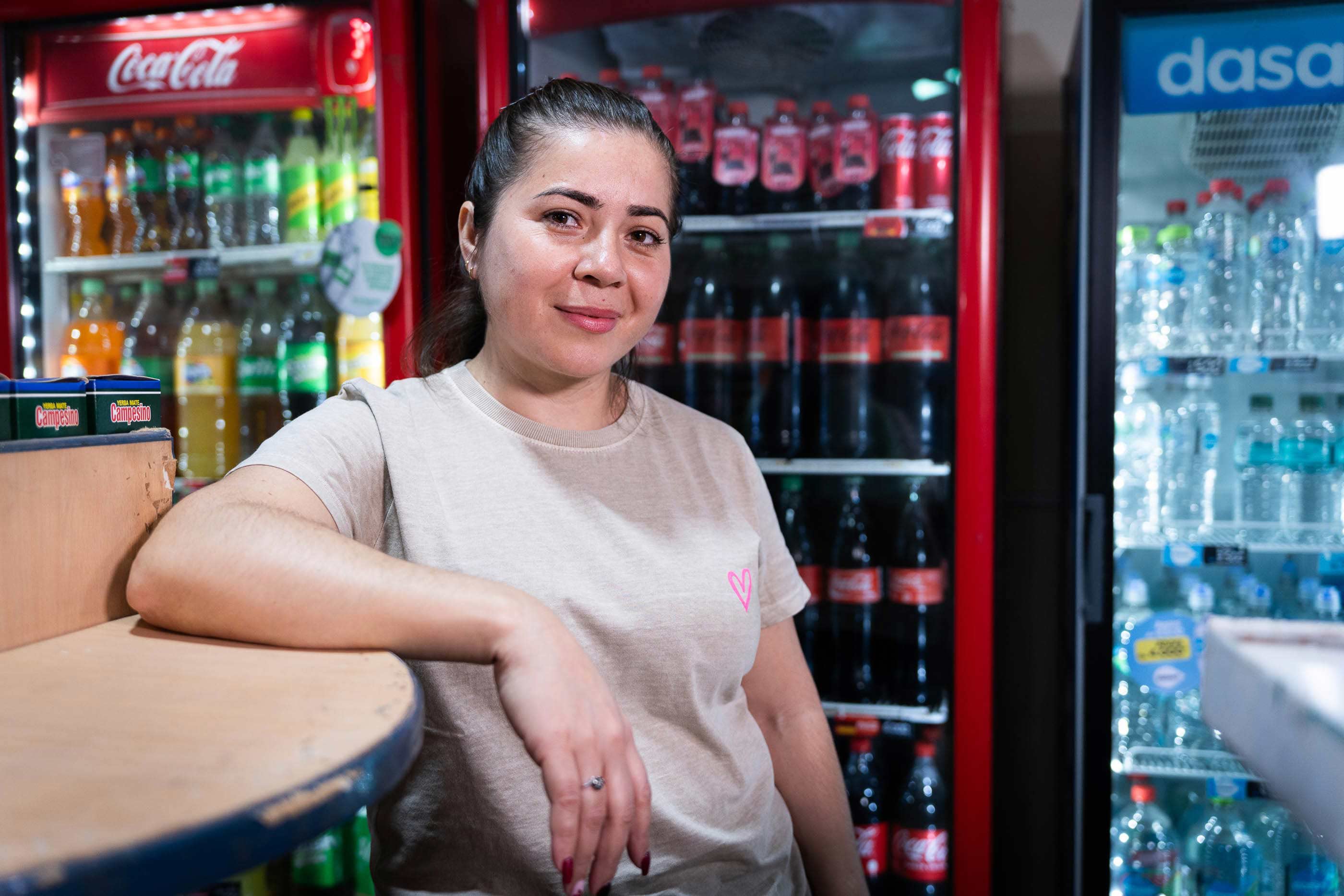 Una mujer apoyada en un mostrador, con una heladera de bebidas al fondo.