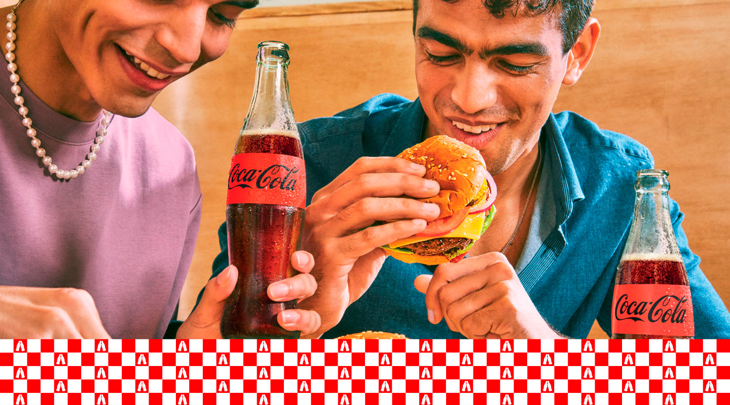 Dos jóvenes sonríen mientras disfrutan de hamburguesas y Coca-Cola en un ambiente relajado.
