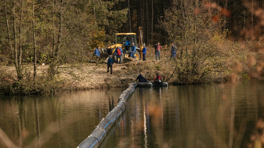 Echipă de muncitori cu excavator pe malul unui râu, pe râu este întinsă o capcană plutitoare pentru oprirea deșeurilor.