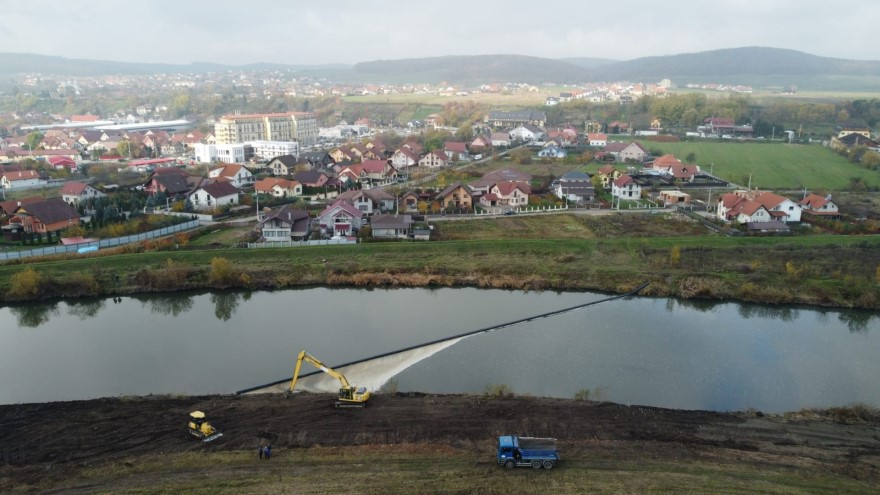 Imagine aeriană cu o capcană plutitoare pentru gunoaiele de pe un râu care trece aproape de un oraș; pe mal se lucrează cu utilaje