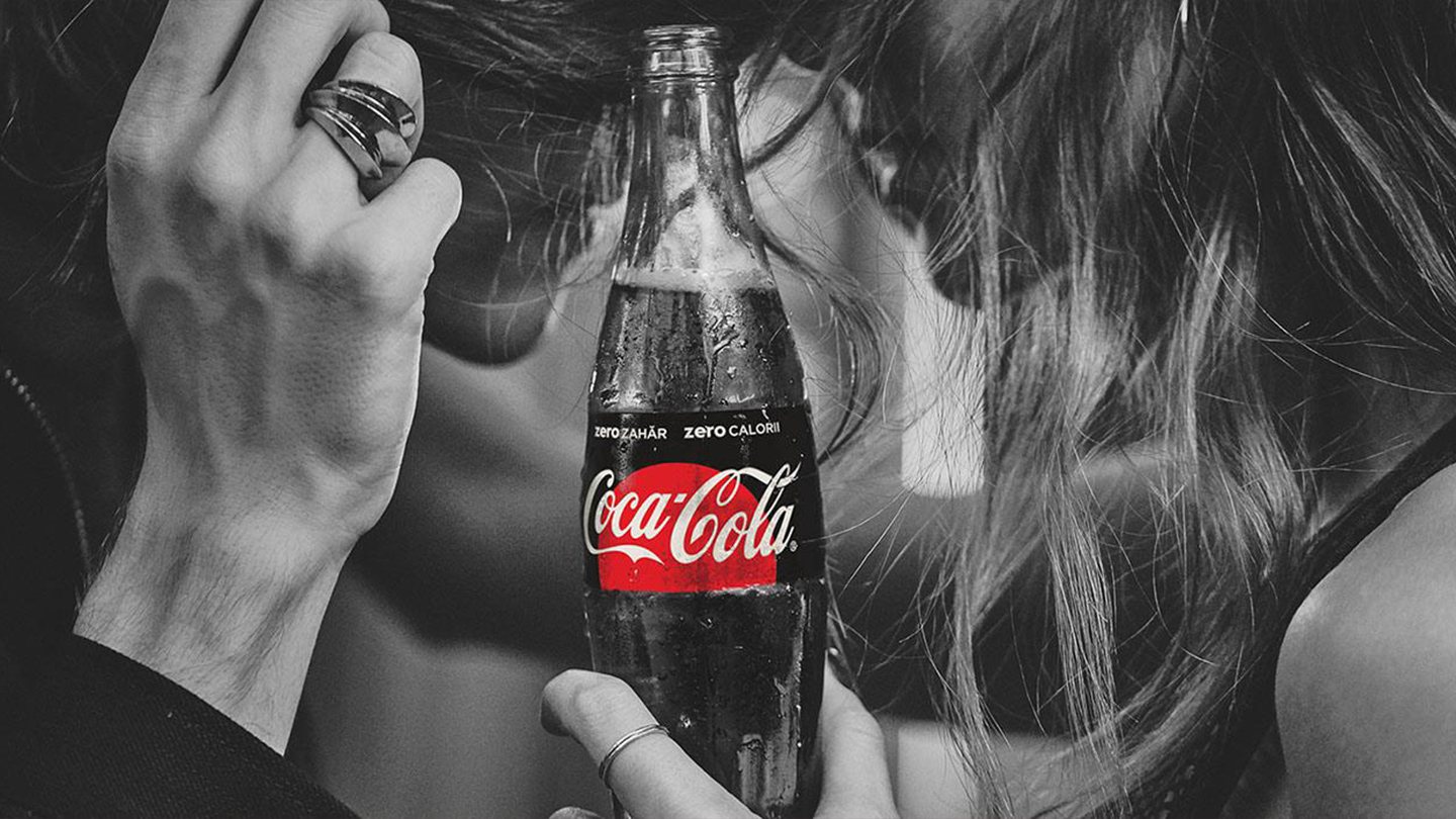 Un tănâr și o tânără, văzuți din profil, îmbrățișați, tânăra ține în mână o sticlă de Coca-Cola Zero Zahăr