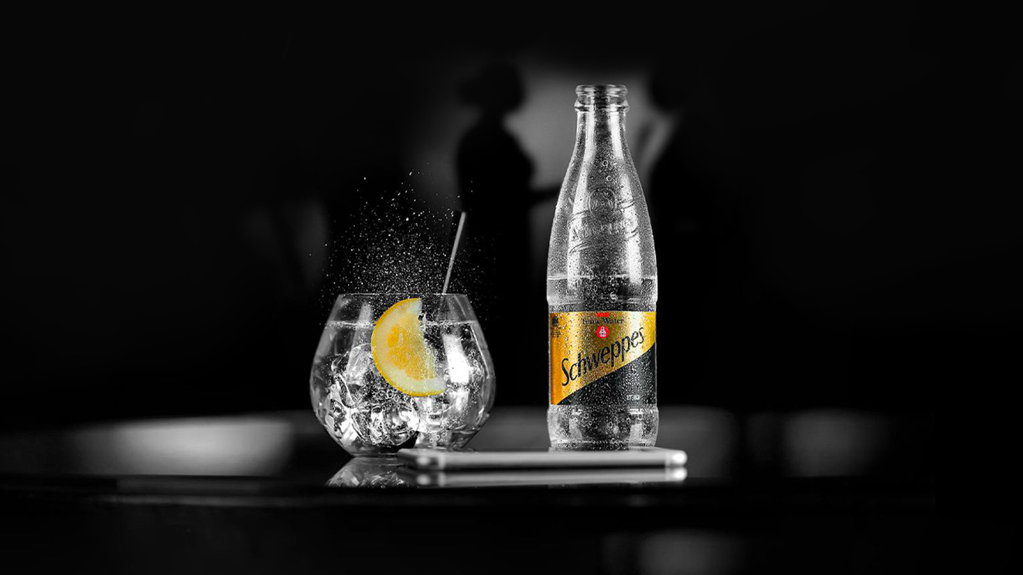 Sticlă și pahar de Schweppes Kinley Tonic, pe fundalul întunecat se văd umbrele unor dansatori.