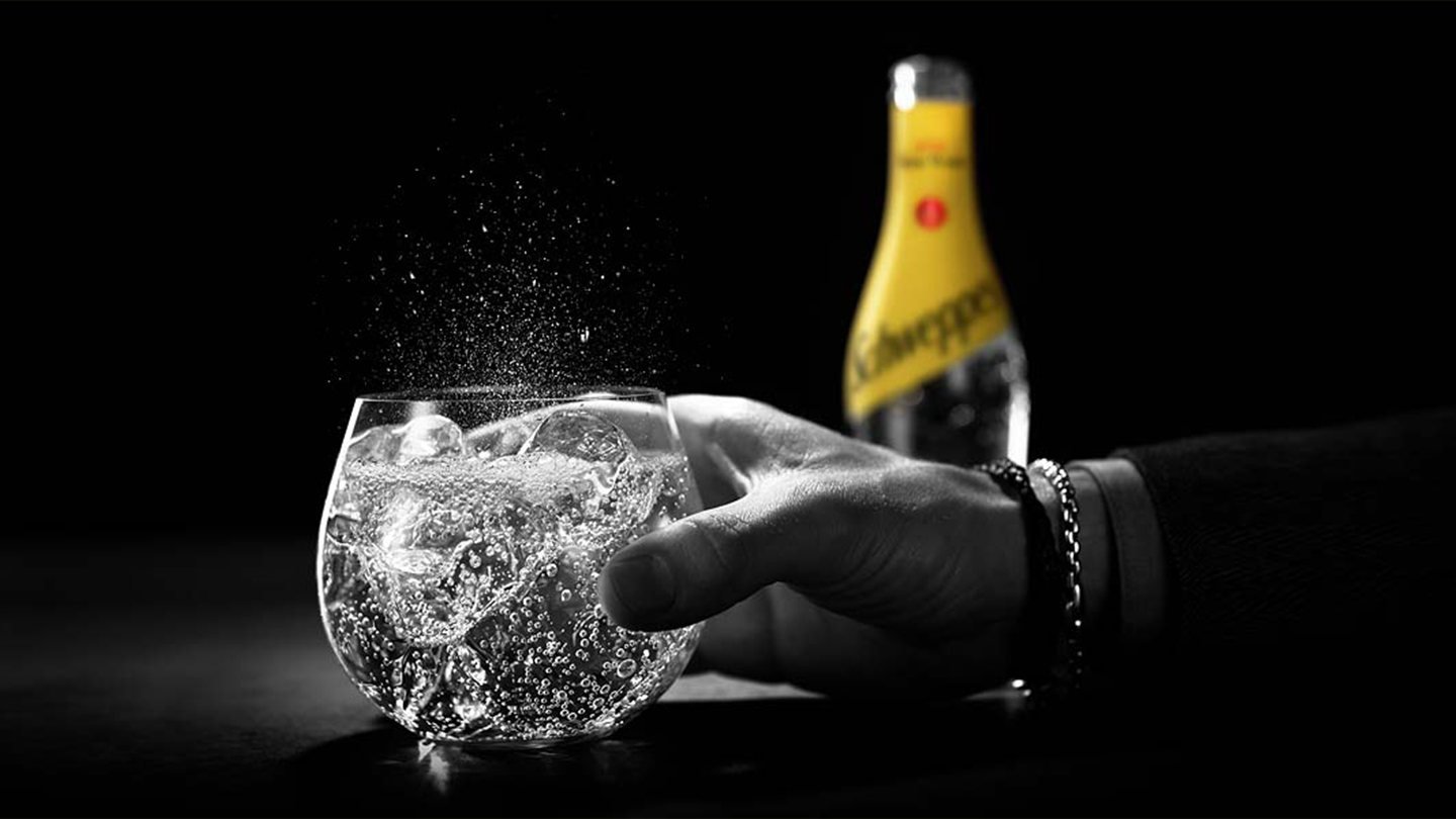 Crno-bela ruka drži čašu Schweppes Tonic water sa Schweppes bocom u pozadini