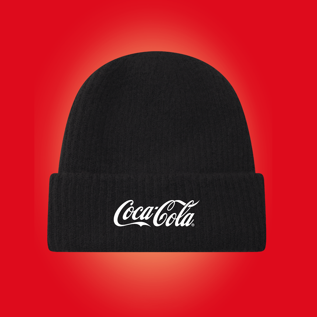 En svart stickad mössa med vit Coca-Cola-logotyp visas mot en röd bakgrund med ljus mittpunkt.