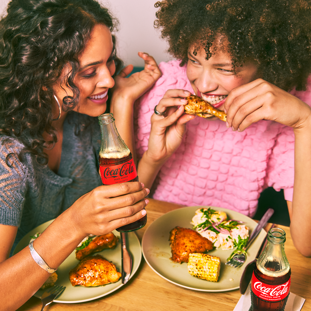 Två kvinnor äter kyckling vid ett bord och dricker Coca-Cola. På tallrikarna finns majs och sallad.