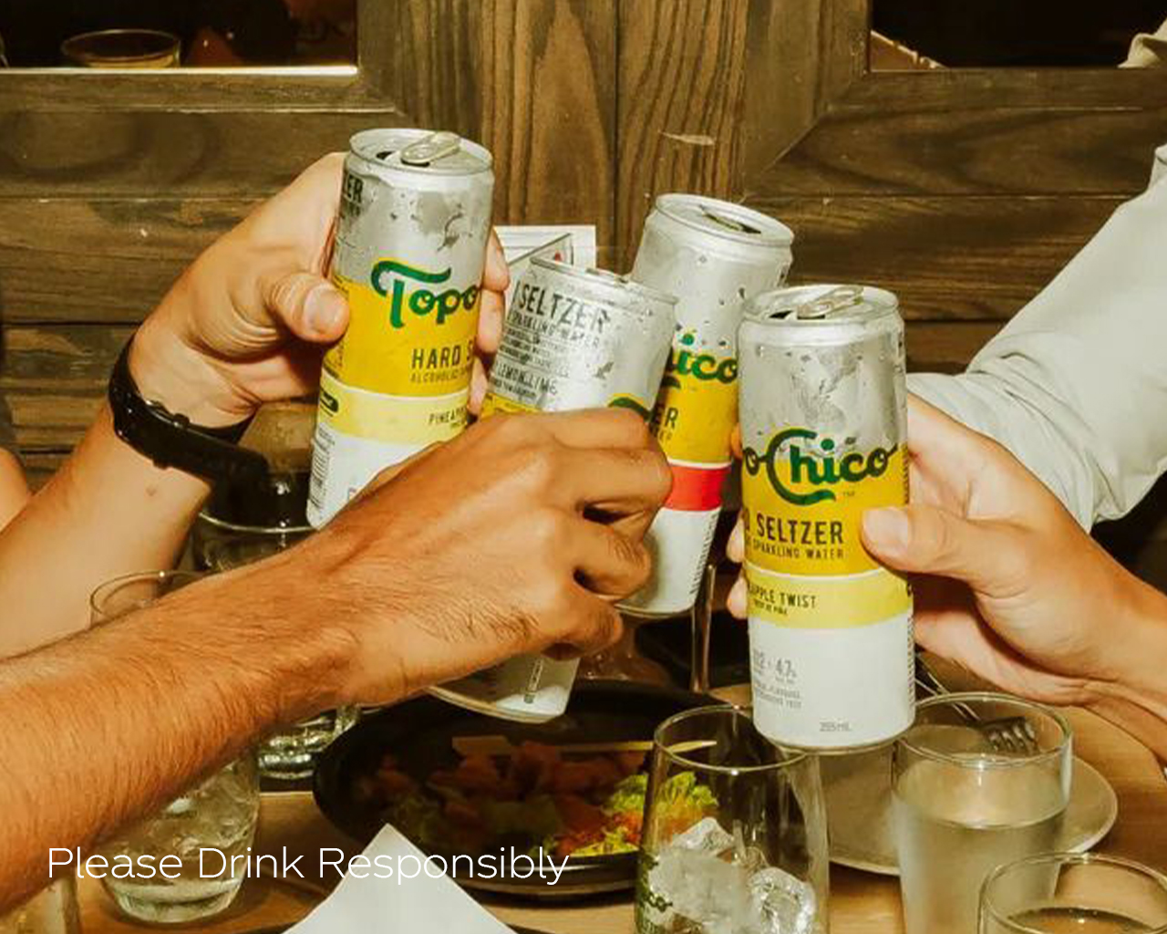 Detail of four arms toasting cans of Topo Chico around a table