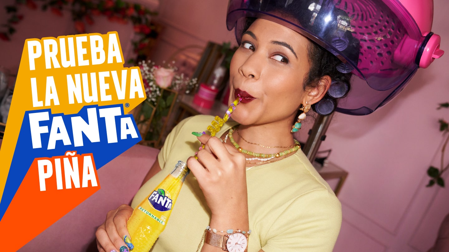 Mujer de pelo enrulado rubio, con anteojos y ropa colorida bebiendo con los ojos cerrados una botella de Fanta sabor piña y un snack en su otra mano dentro de una biblioteca. A su lado hay un texto que dice “El match perfecto con tu snack favorito ¡Si existe!” y por debajo está el logo de Fanta.