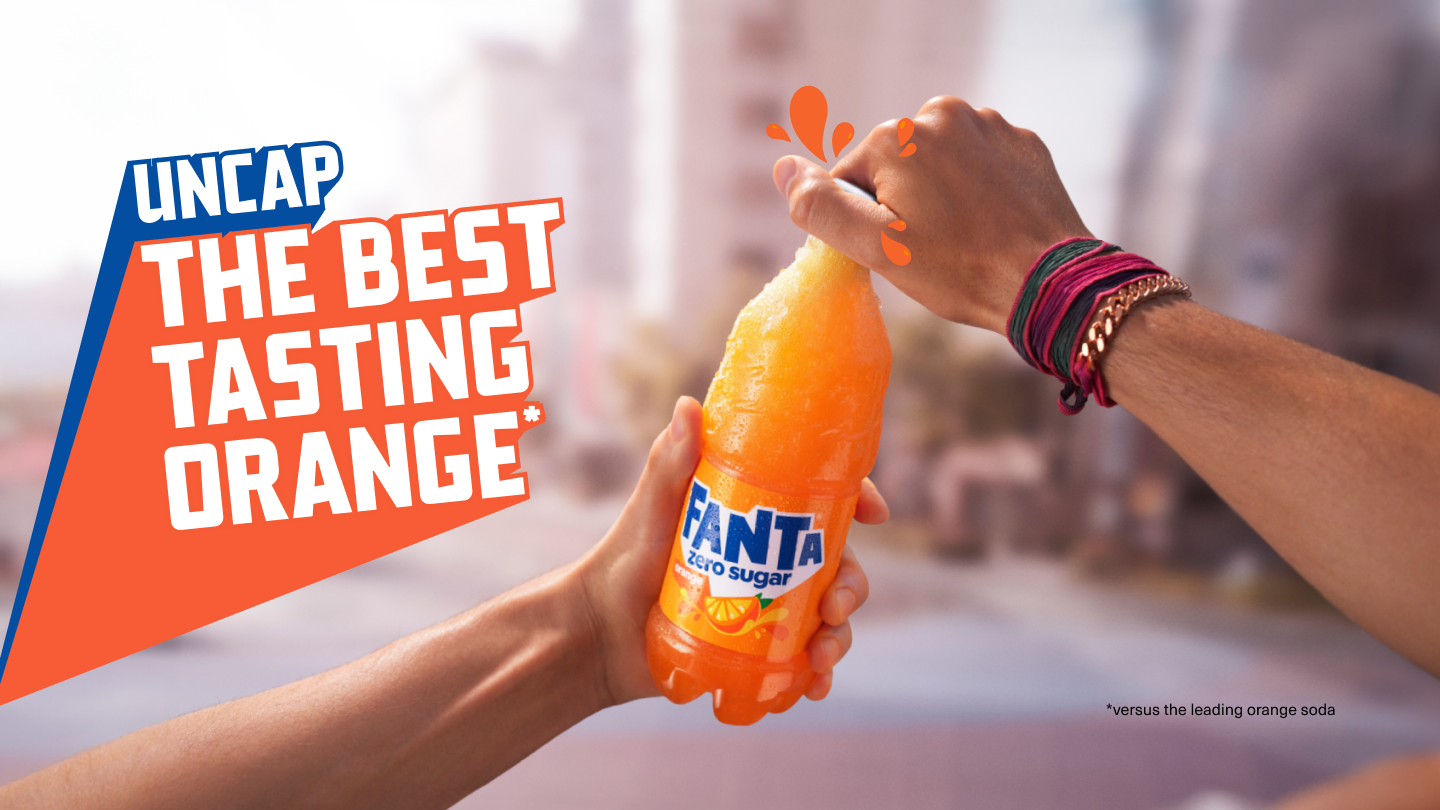 A hand holding a bottle of Fanta Orange and in the background a group of friends smiling and drinking Fanta