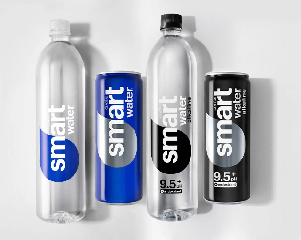 Four Glacéau Smartwater products laid flat side by side on a white background: a bottle and can of original Smartwater with blue labels, and a bottle and can of Smartwater Alkaline with black labels marked "9.5+ pH" and "antioxidant."