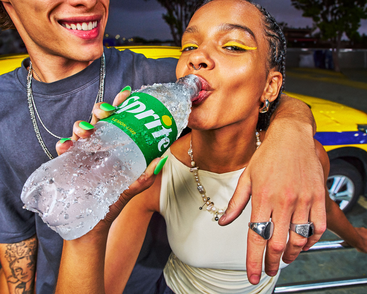 A smiling friend leans on a woman as she drinks from a bottle of Sprite in front of a yellow car.