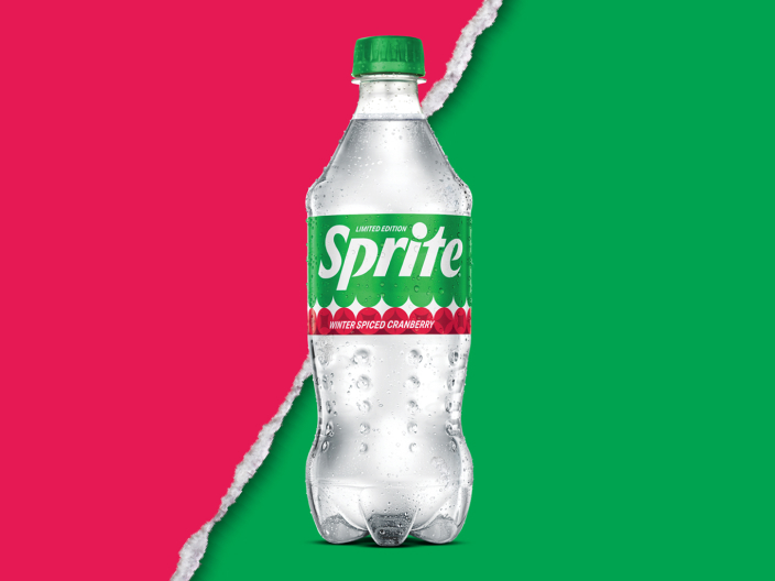 A clear plastic bottle of Sprite Winter Spiced Cranberry with a green cap. It has a green “Limited Edition” label with a red and white scalloped band, and it sits against a background split diagonally between pink and green by a torn-paper edge.