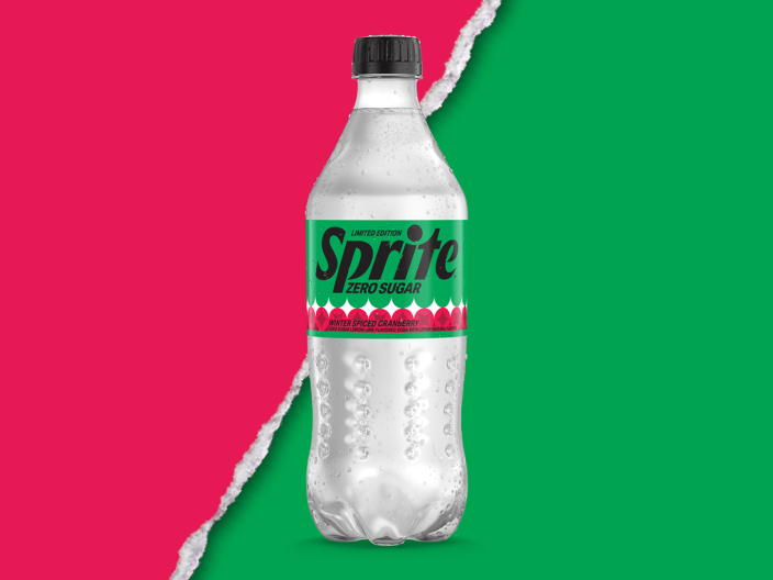 A clear plastic bottle of Sprite Zero Sugar Winter Spiced Cranberry with a black cap. It has a green “Limited Edition” label with a red and white scalloped band, and it sits against a background split diagonally between pink and green by a torn-paper edge.
