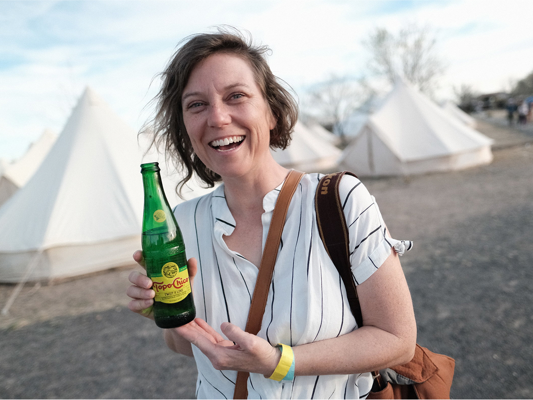 A smiling woman holding a bottle of topo chico at a campsite