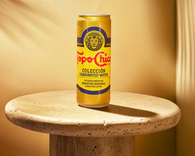 A gold can of Topo Chico Colección Carbonated Water sits atop a round stone pedestal table, lit with warm diagonal light streaks against a tan wall.
