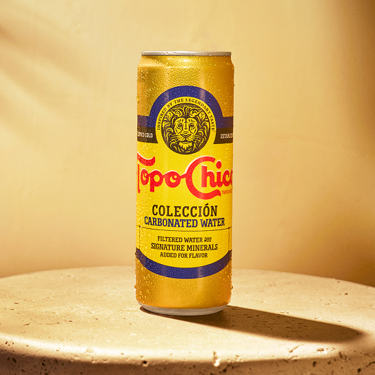 A gold can of Topo Chico Colección Carbonated Water, beaded with condensation, stands on a round travertine table against a warm tan backdrop with soft diagonal light streaks. The label reads "Filtered Water & Signature Minerals Added for Flavor."