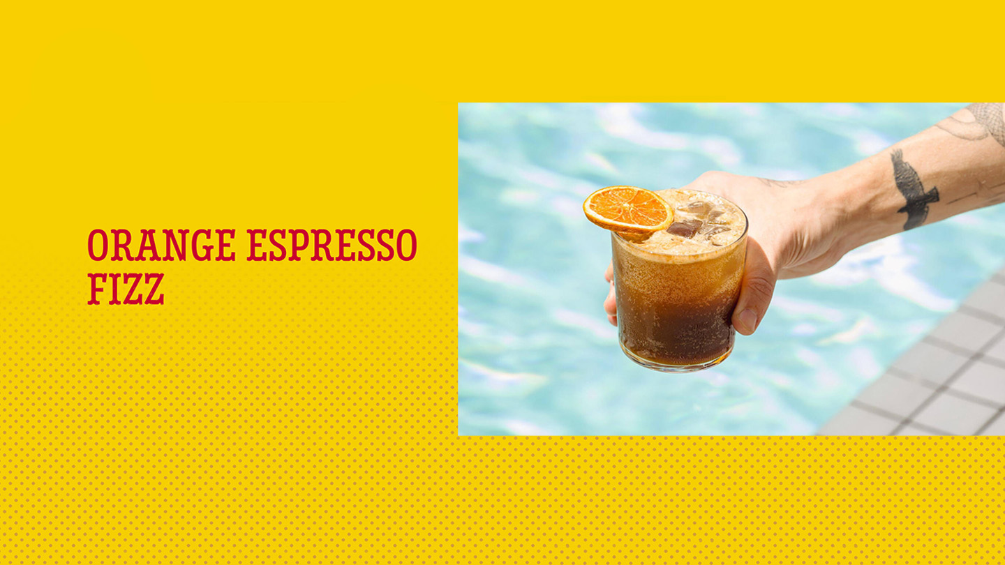 Detail of a hand holding a glass of Orange Espresso Fizz drink with a pool on the background