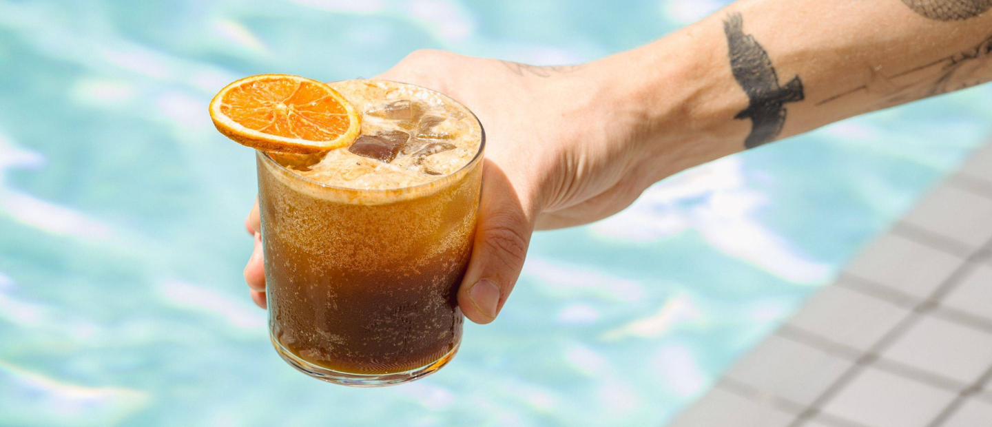 Detail of a hand holding a glass of Orange Espresso Fizz drink with a pool on the background
