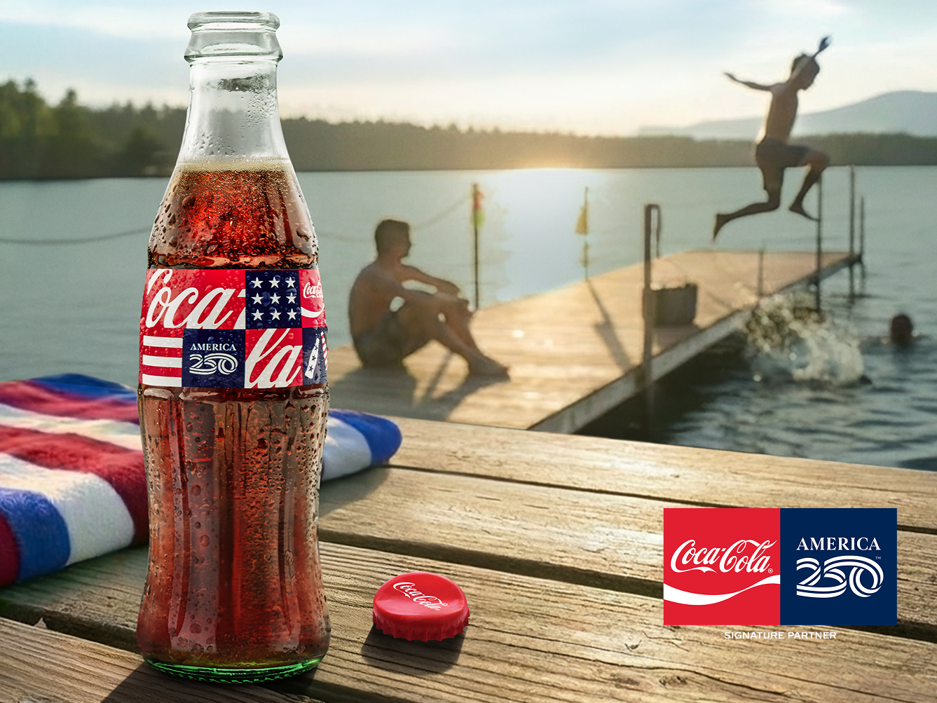 Chilled Coca‑Cola glass bottle with America250 label on lakeside dock as friends jump into the water at sunset