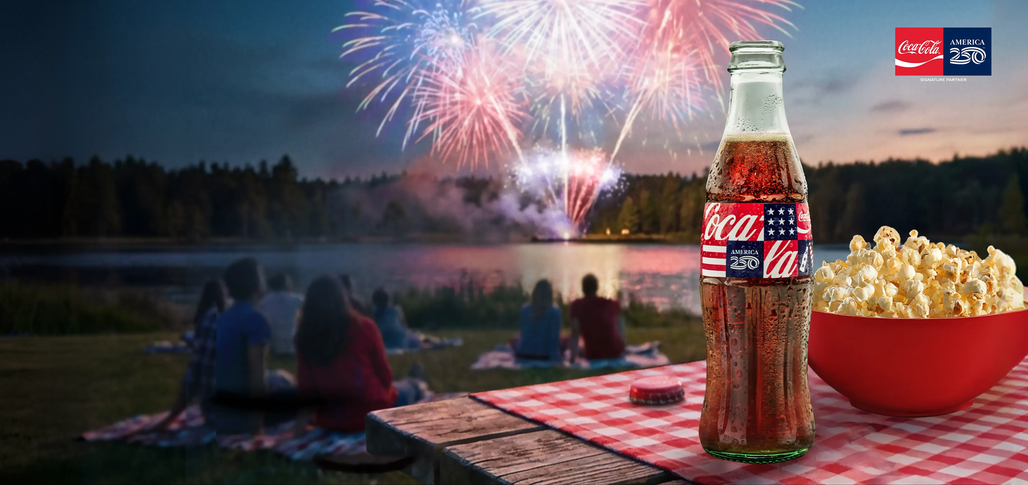 Glass Coca‑Cola bottle with America250 flag label and popcorn on picnic table as people watch fireworks over a lake.