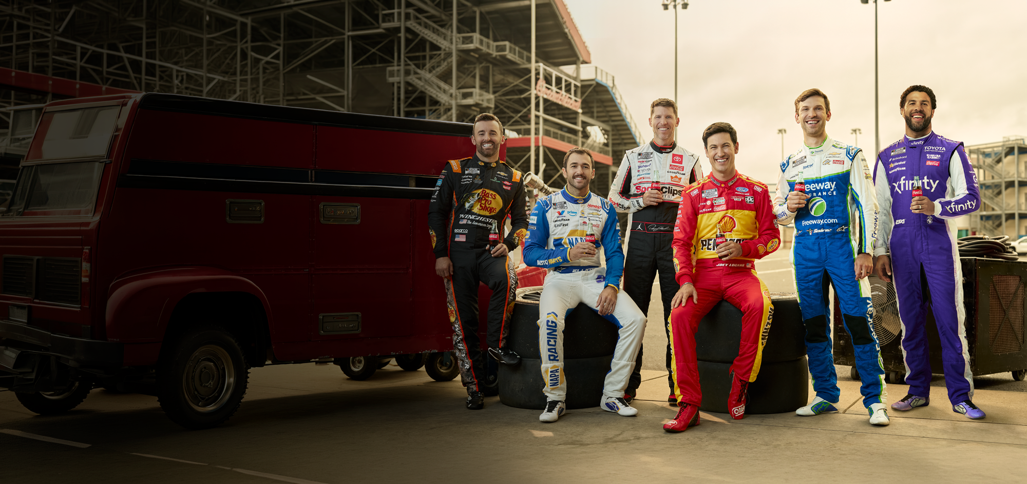 Six NASCAR drivers in racing suits hold Coca-Cola bottles at the track for a Daytona 500 promotional banner.