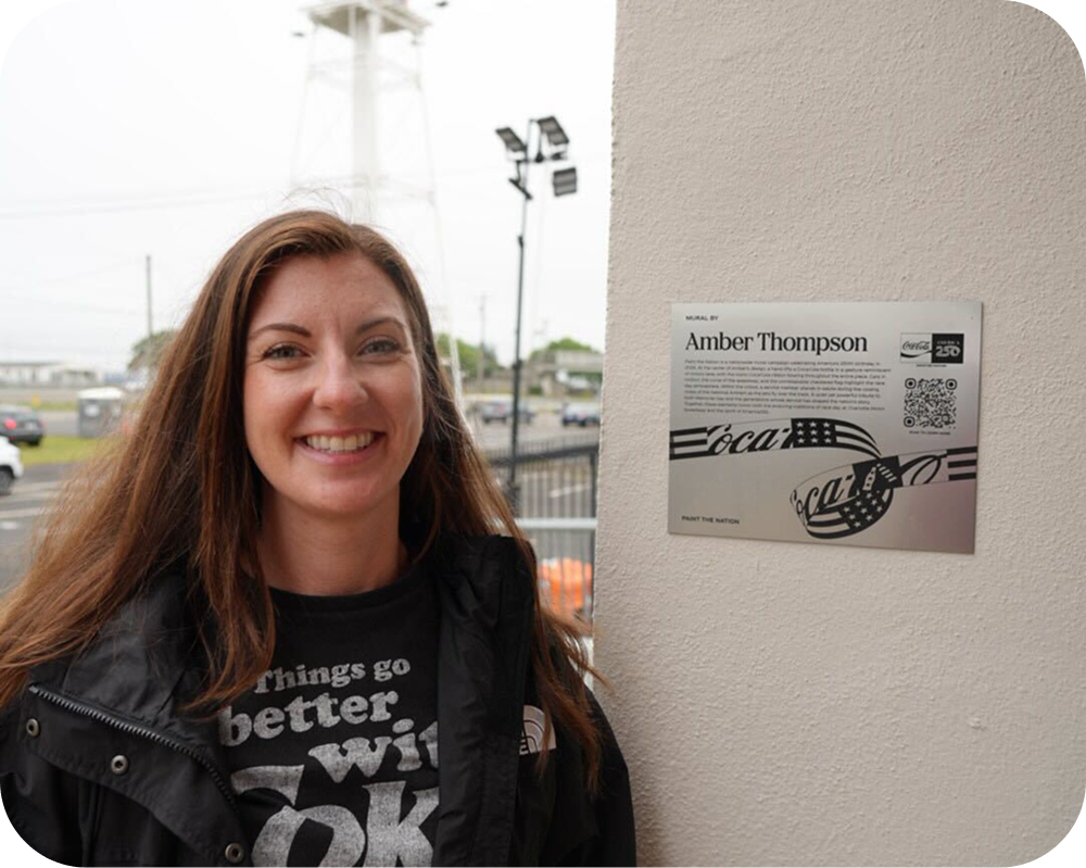 Artist Amber Thompson smiling next to her Paint the Nation exhibit plaque for the Coca‑Cola mural.