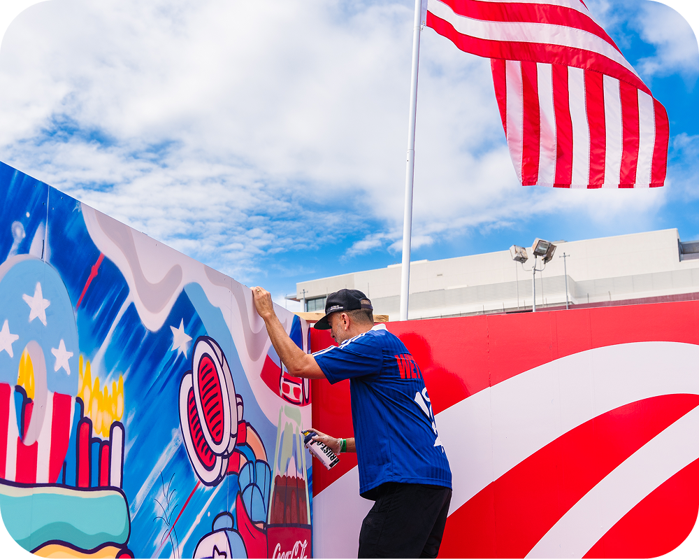Dave Ross spray painting his America 250 Coca‑Cola mural outdoors with an American flag flying in the background.