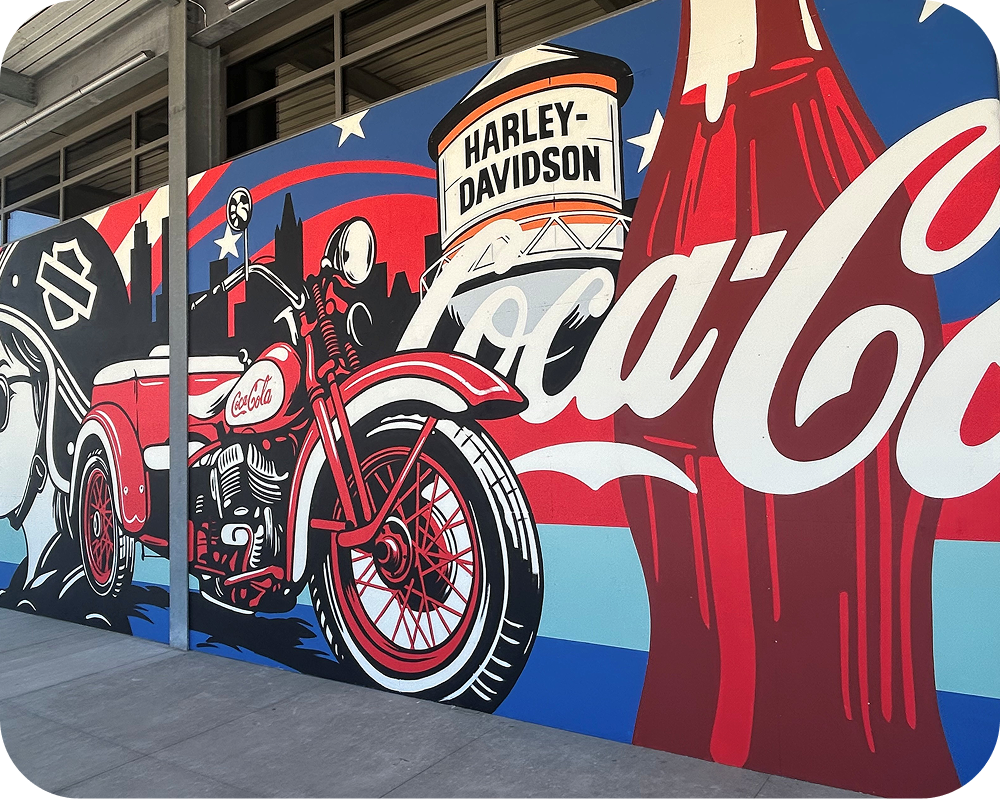 Section of the Greg Gossel mural displaying a red motorcycle, Harley-Davidson water tower, and large Coca‑Cola logo.