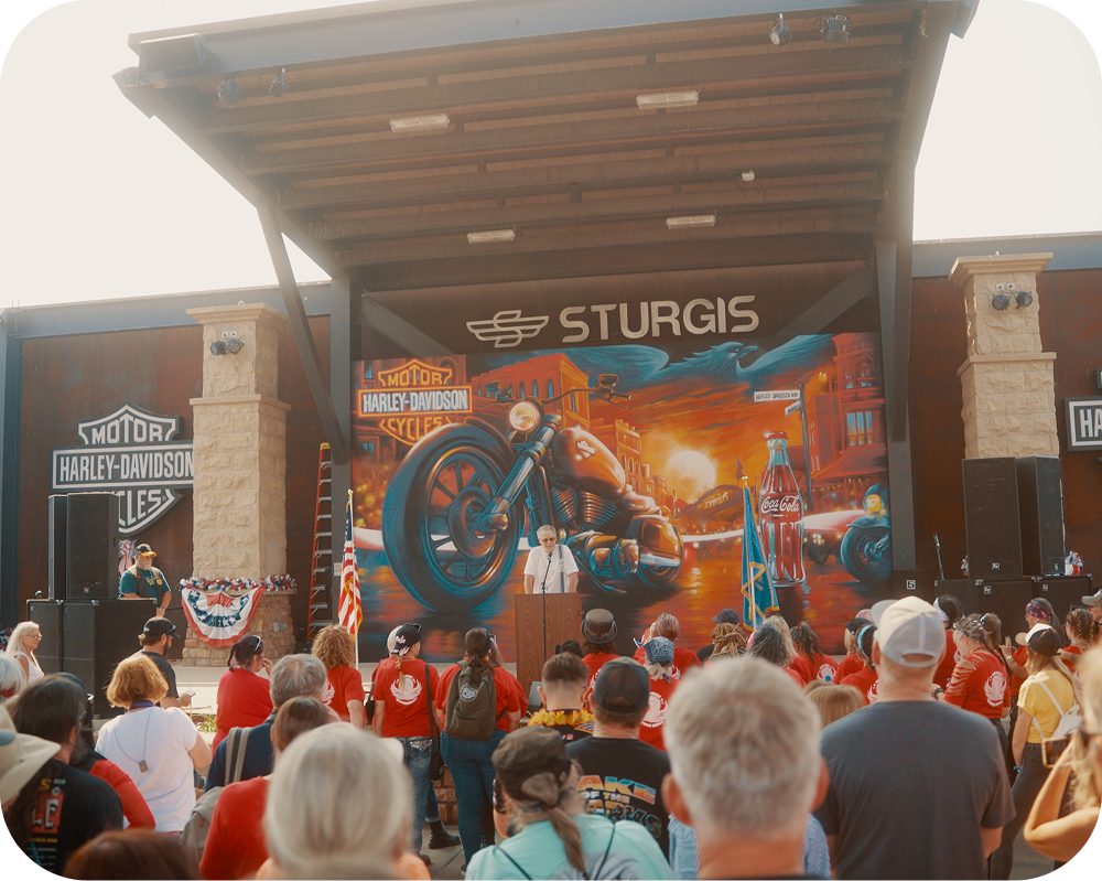 A crowd watches a speaker at a podium on a stage featuring the Sturgis America 250 Coca-Cola mural backdrop.