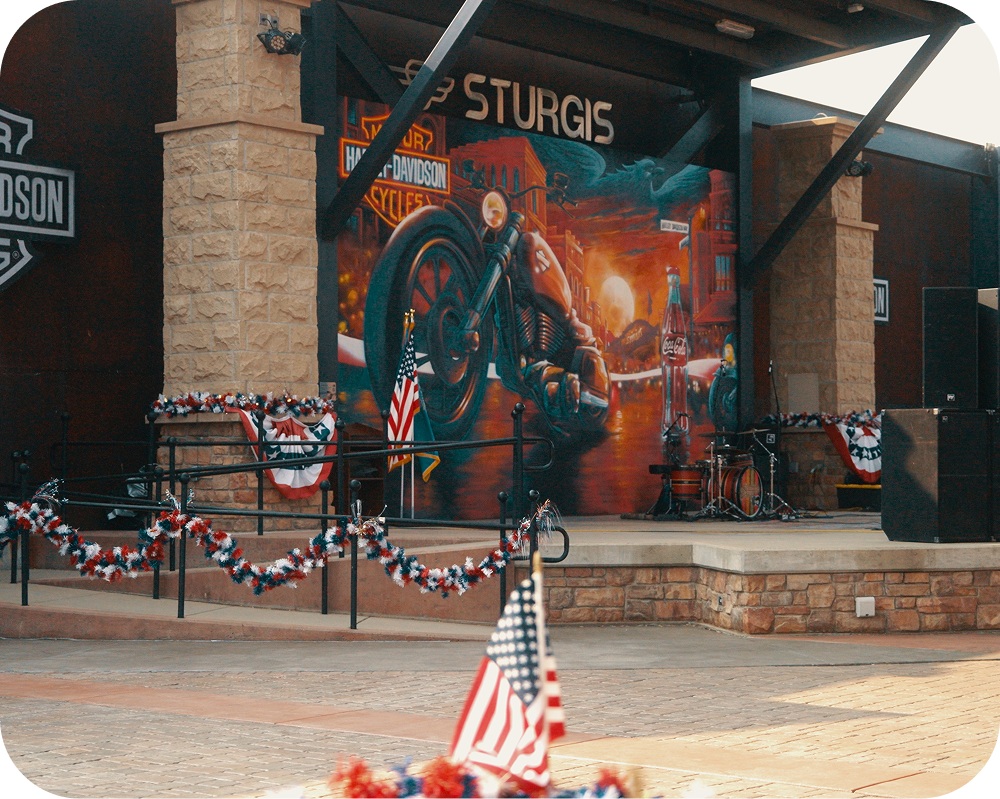 Outdoor stage decorated with patriotic bunting and flags framing the Sturgis America 250 Coca-Cola mural.