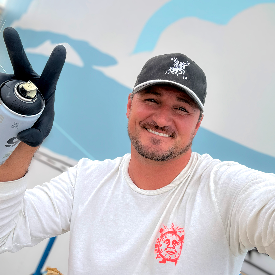 Mural artist Jordan Dean holding a spray paint can and making a peace sign while working on his America 250 mural.