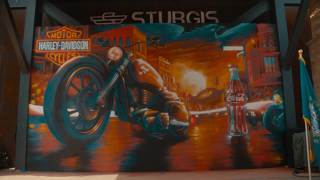 America 250 mural by Jordan Dean in Sturgis featuring a Harley-Davidson motorcycle and a glass Coca-Cola bottle.