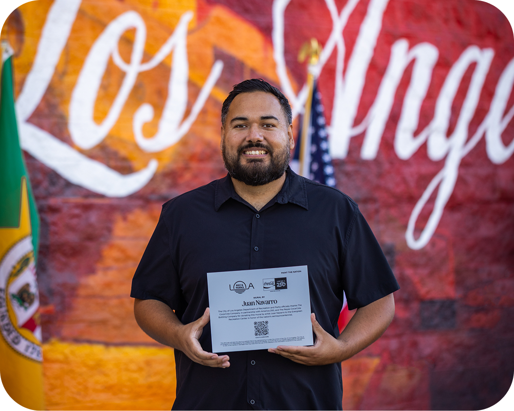 Mural artist Juan Navarro holding an America 250 dedication plaque in front of his Los Angeles Coca-Cola artwork.