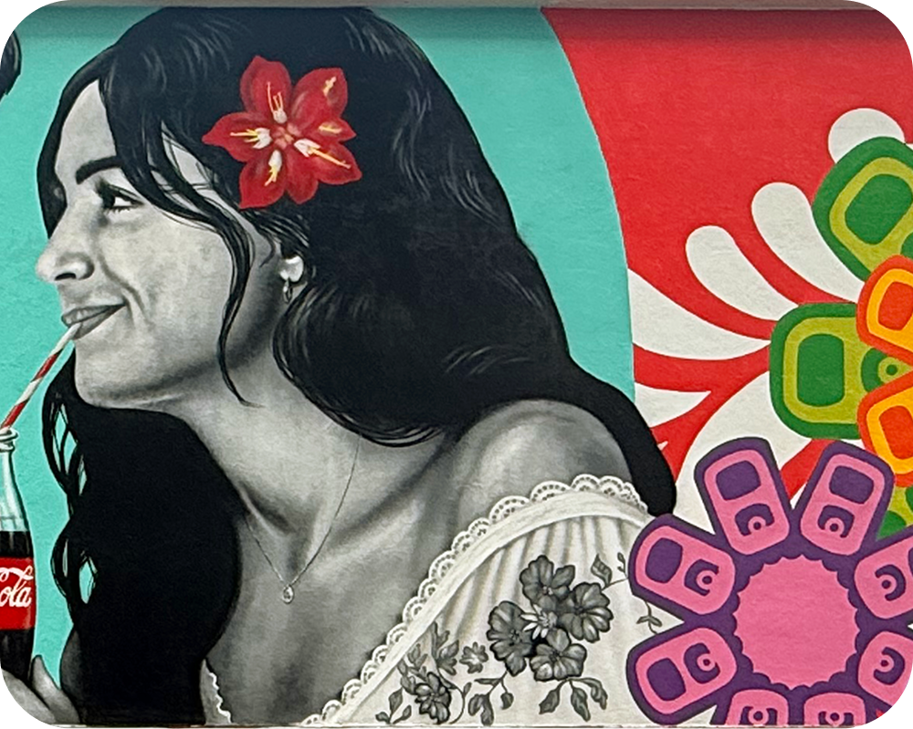 Smiling woman with a red flower in her hair drinking from a Coca‑Cola bottle in the Los Otros mural.