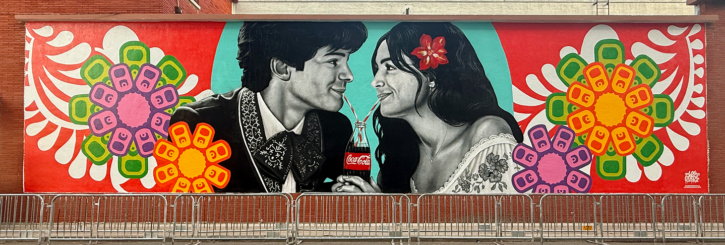 Vibrant 'Los Otros' street art mural on a brick wall, featuring a couple in traditional attire sharing a glass Coca-Cola bottle.