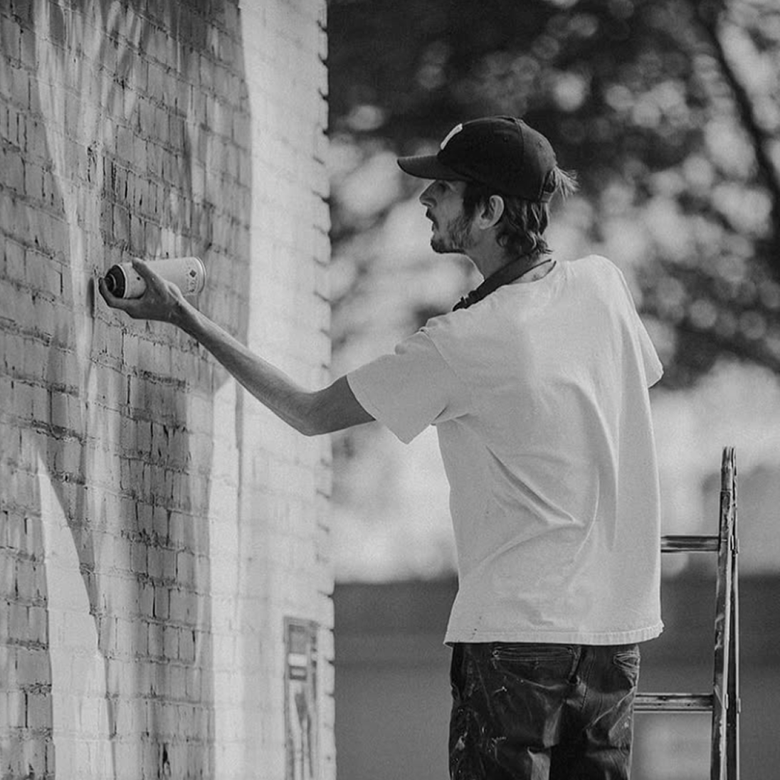 Artist Malcolm Byers spray-painting a brick wall while working on his America 250 Coca-Cola mural project.
