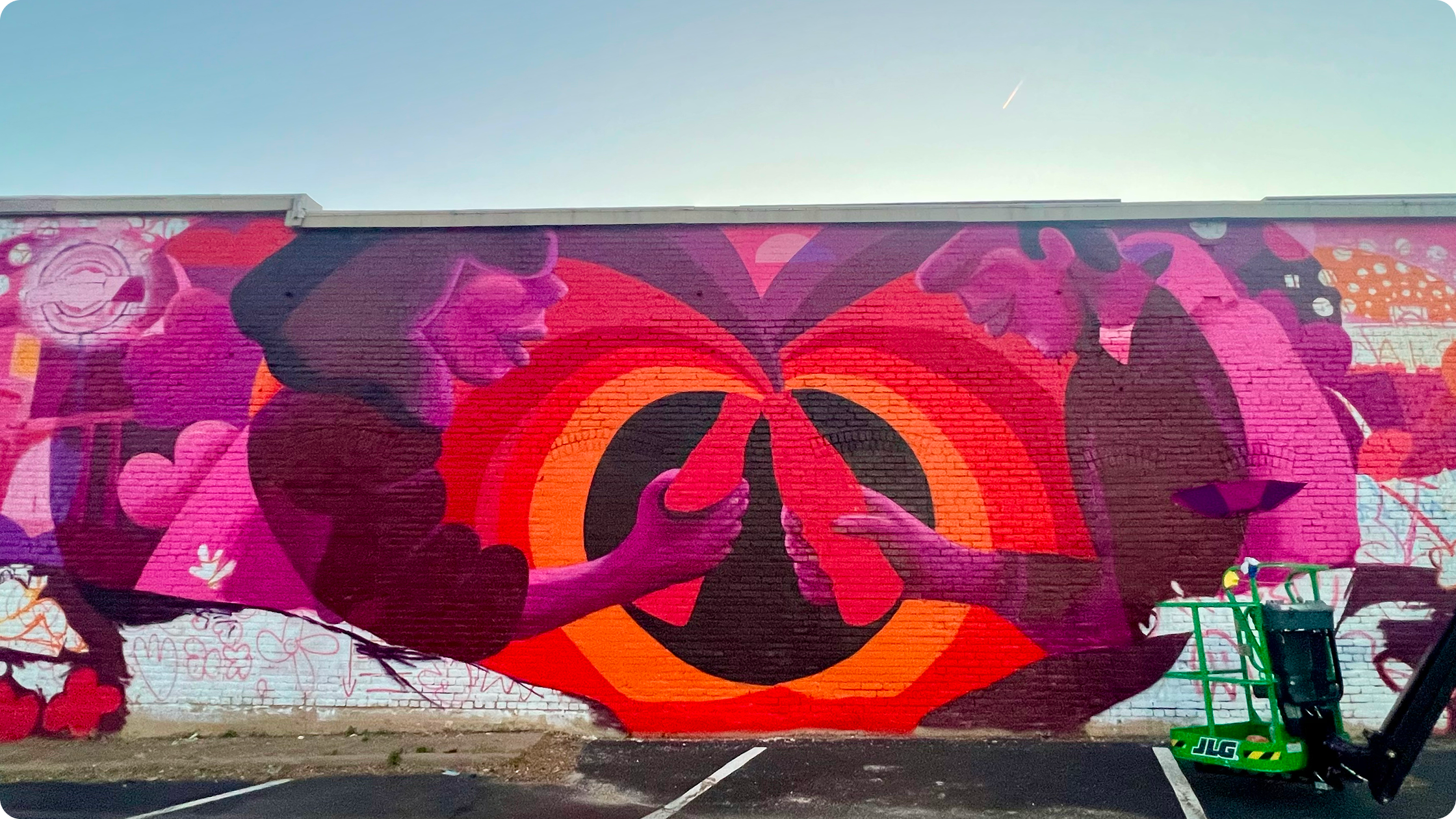 Wide brick wall mural by Meg Wagler with two stylized figures clinking Coca-Cola bottles in bold pink, red and orange tones.