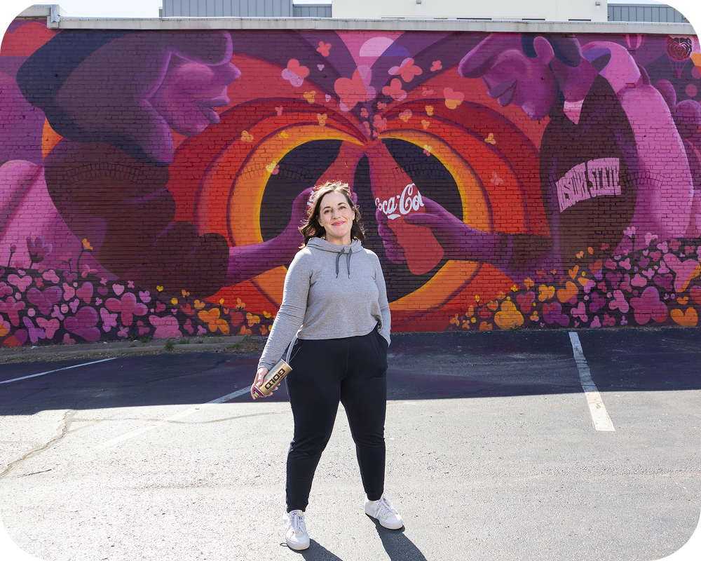 Artist posing in front of the finished Paint the Nation Coca-Cola mural, standing in parking lot with colorful hearts behind