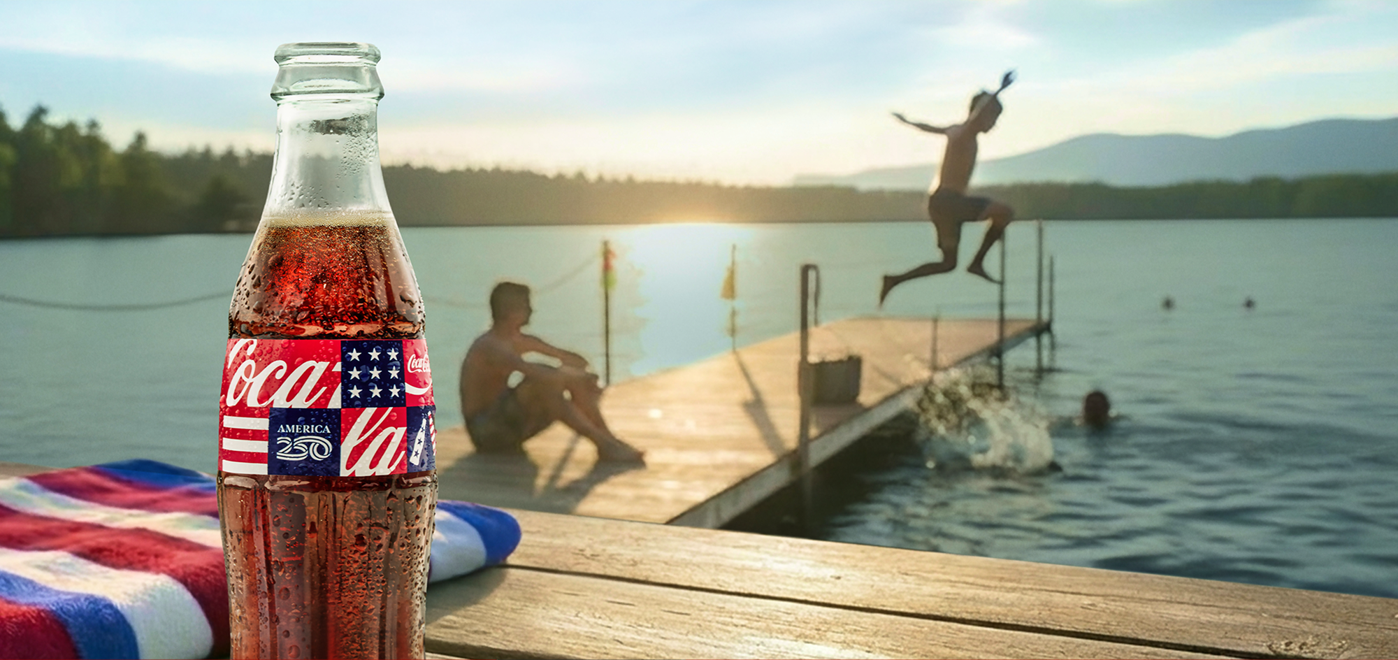 Coca-Cola glass bottle with an America250 label sits on a wooden dock while people swim and jump into a sunny lake.