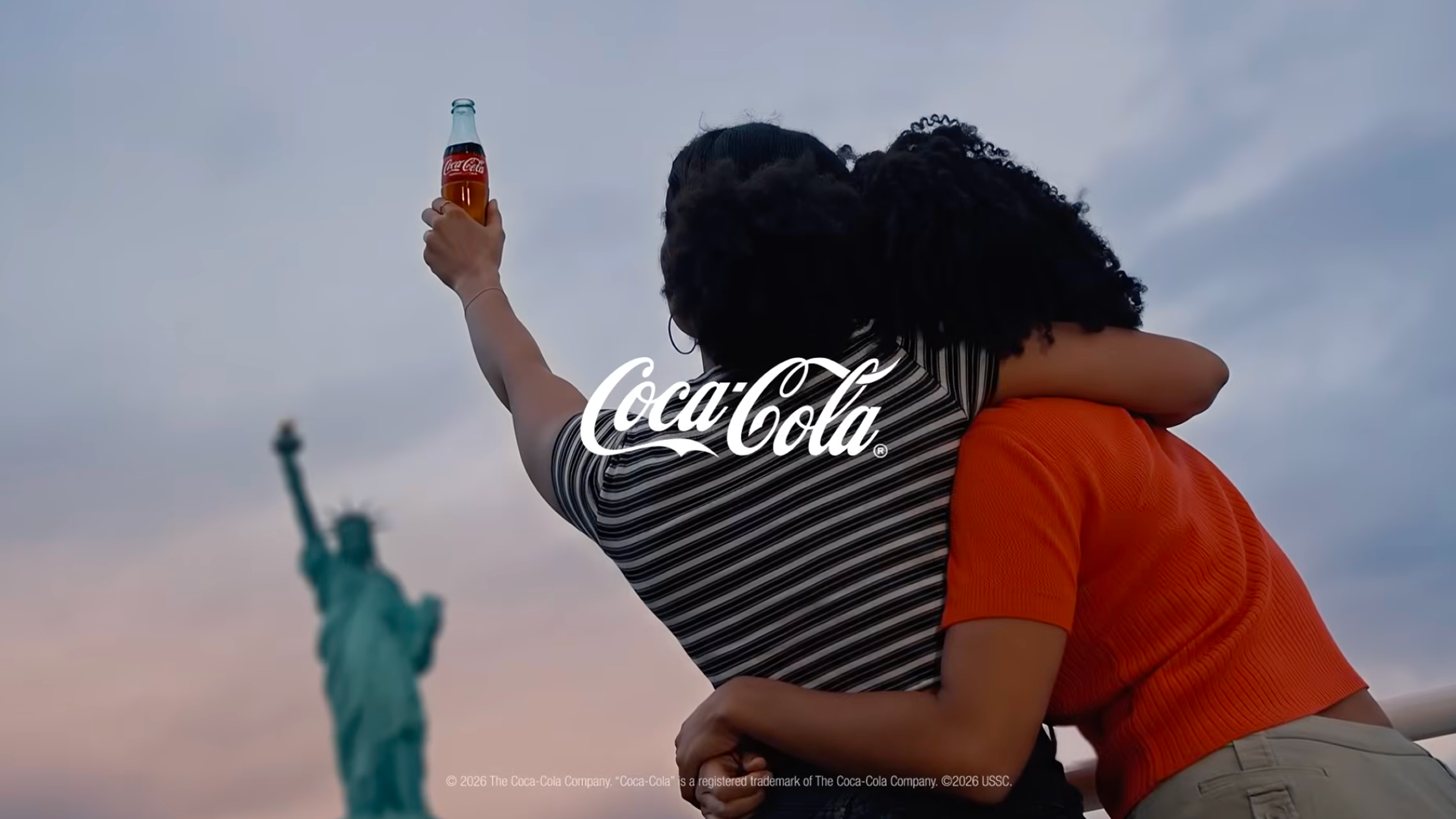 Two friends hug while holding up a Coca-Cola glass bottle toward the Statue of Liberty during a beautiful sunset.