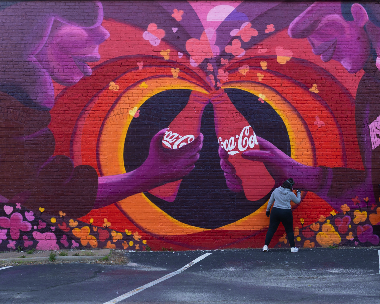 Artist paints a vibrant brick wall mural featuring two hands toasting Coca-Cola glass bottles inside a bright heart.