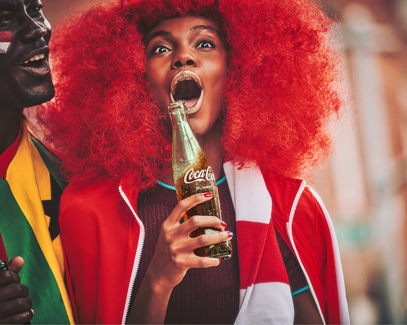 Fan with bright red curly hair holding icy Coca-Cola glass bottle while celebrating match day in the street.